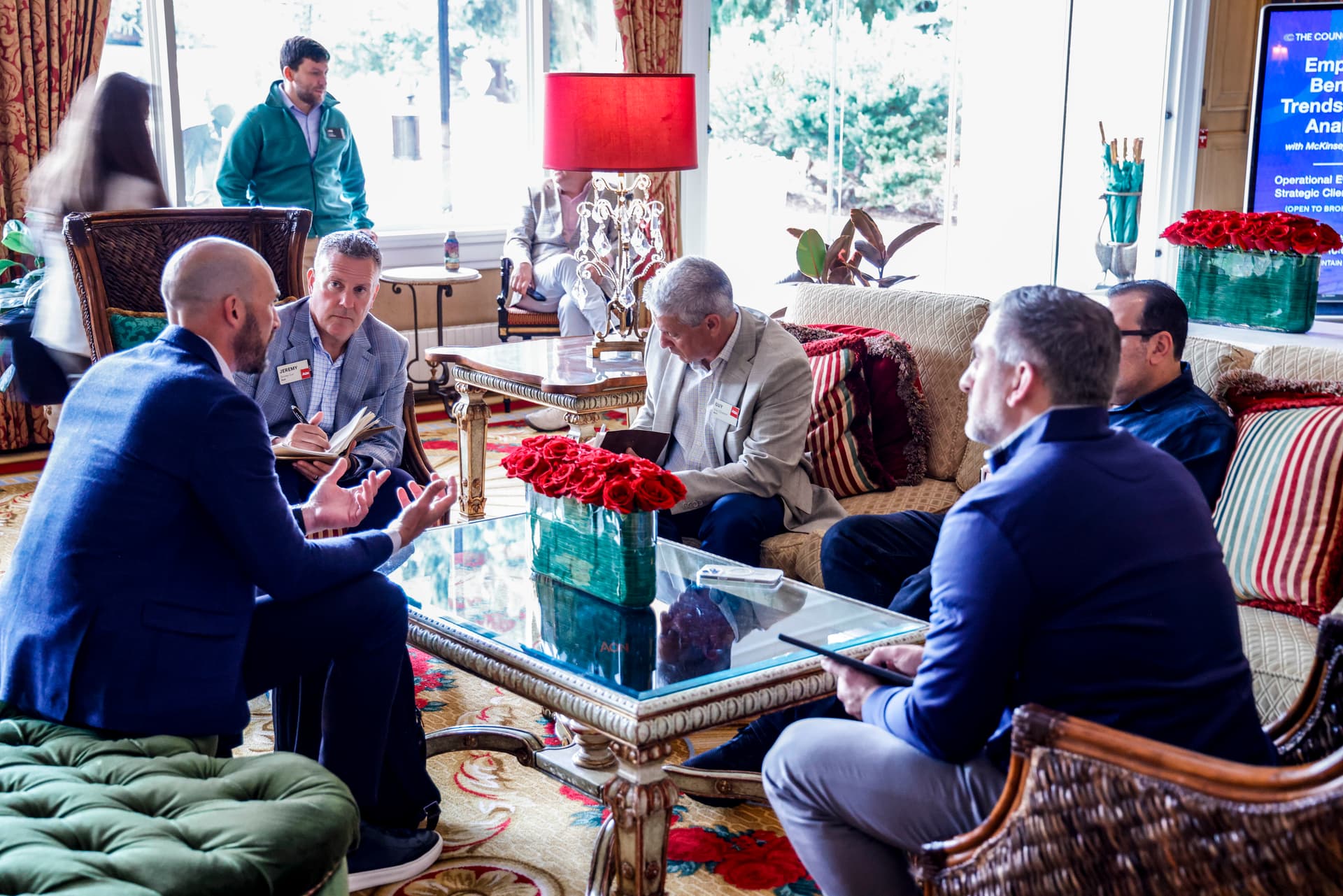A small group of business professionals engaged in a collaborative discussion during a networking or leadership event. Participants sit around a coffee table in a comfortable lounge setting, exchanging ideas and taking notes while natural light fills the room. Floral arrangements, upholstered seating, and a presentation screen in the background create a professional yet conversational atmosphere.